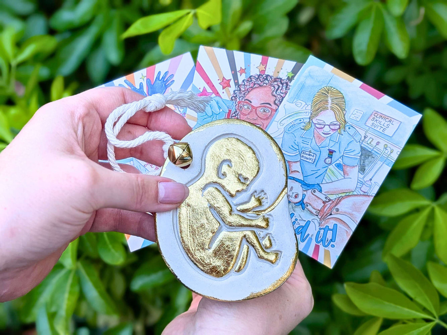 A hand holds a round white ornament featuring a raised gold fetus design and a small gold bell, with two illustrated cards behind it against a background of green leaves.