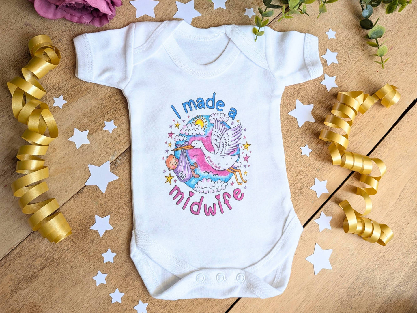 A white short-sleeve baby bodysuit laid flat on a wooden surface, decorated with a colorful graphic of a stork and the text 'I made a midwife.' Gold ribbon curls, white star cutouts, and greenery are arranged around it as styling props.