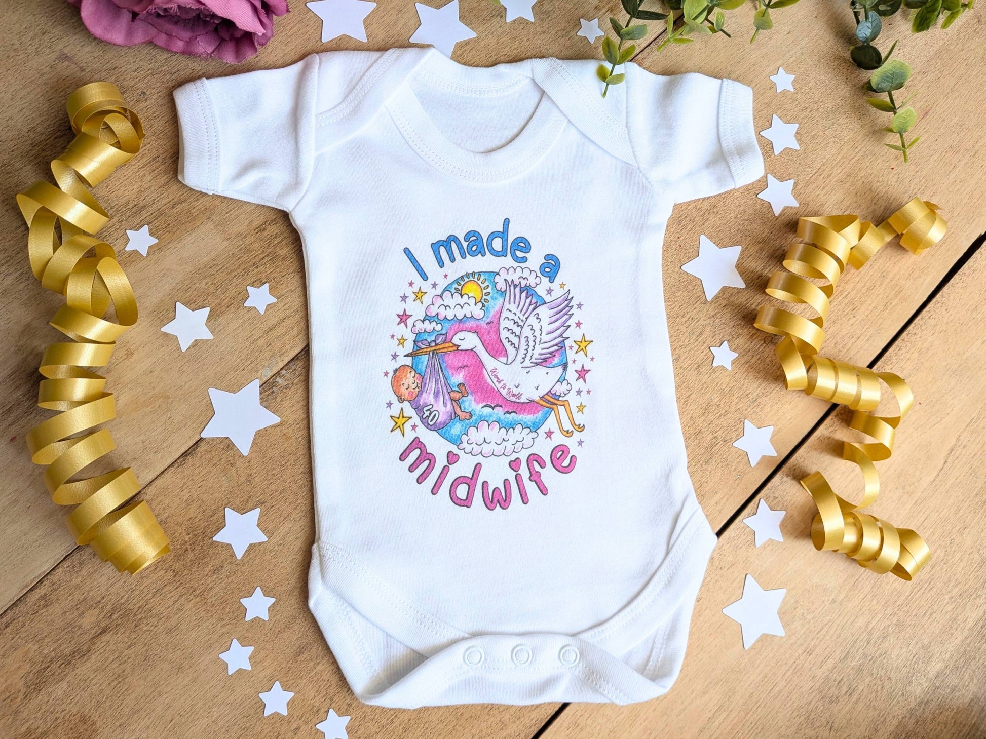 A white short-sleeve baby bodysuit laid flat on a wooden surface, decorated with a colorful graphic of a stork and the text 'I made a midwife.' Gold ribbon curls, white star cutouts, and greenery are arranged around it as styling props.