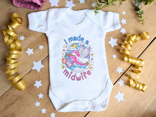 A white short-sleeve baby bodysuit laid flat on a wooden surface, decorated with a colorful graphic of a stork and the text 'I made a midwife.' Gold ribbon curls, white star cutouts, and greenery are arranged around it as styling props.