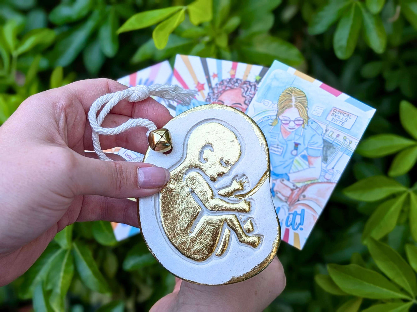 A hand holds a white oval ornament with a gold fetus design and a white hanging cord, with two colorful art cards behind it against a background of green leaves.