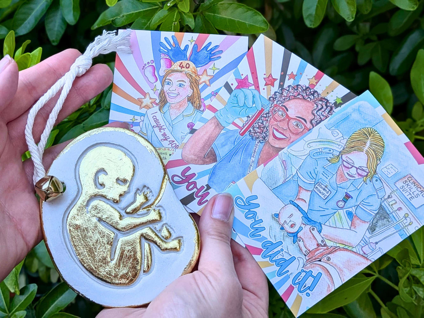 A hand holds a white and gold fetus-shaped ornament with a white rope loop, alongside three colorful illustrated cards featuring midwife art and text, set against a background of green foliage.