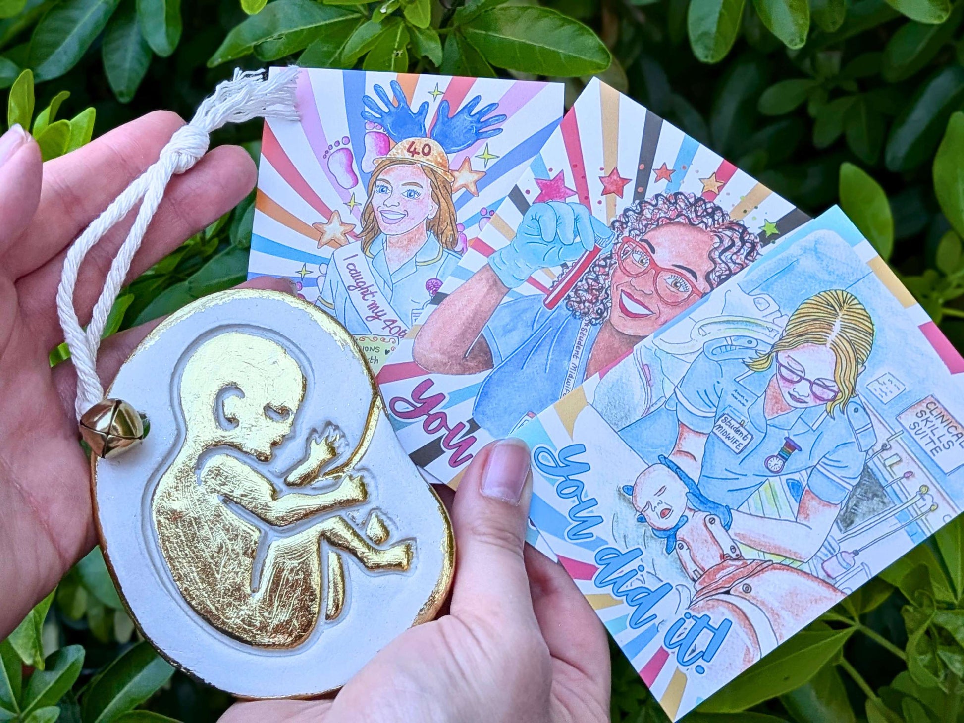 A hand holds a white and gold fetus-shaped ornament with a white rope loop, alongside three colorful illustrated cards featuring midwife art and text, set against a background of green foliage.
