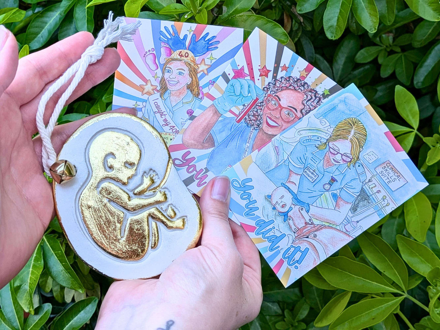 A hand holds a white oval ornament with a gold fetus design and a white cord, while another hand holds three illustrated cards with colorful medical-themed drawings, set against green foliage.