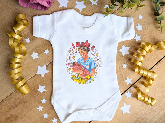 A white short-sleeve baby bodysuit laid flat on a wooden surface, decorated with gold ribbon curls, white star cutouts, and greenery. The bodysuit features a colorful graphic of a midwife holding a baby with the text 'I made a midwife'.