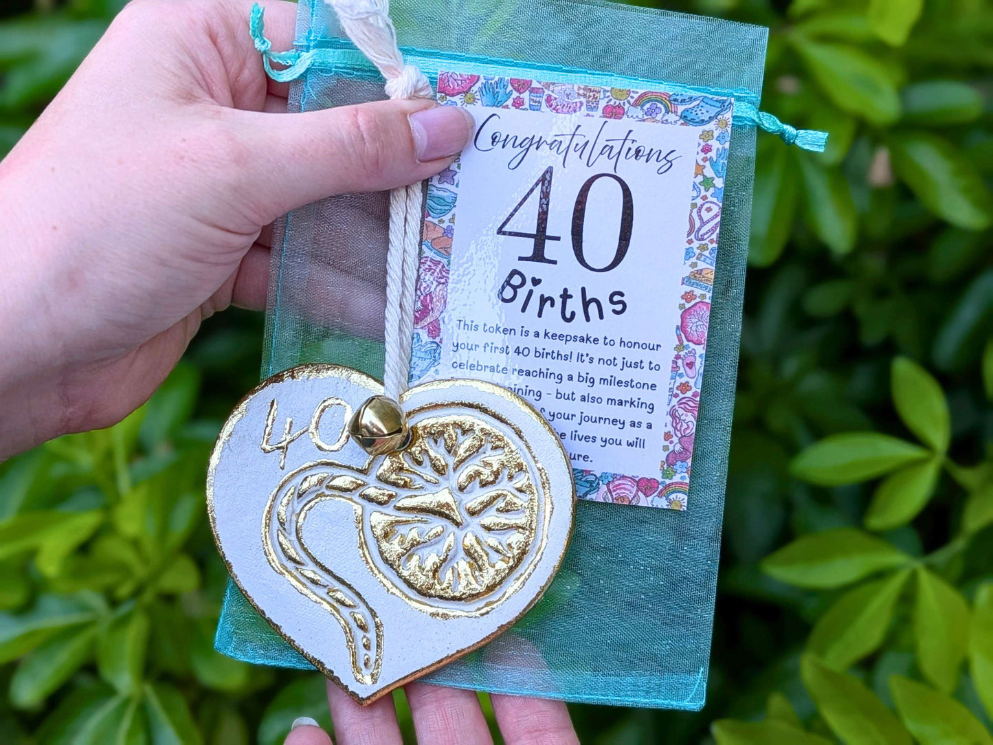 A hand holding a white heart-shaped wooden 40th birth student midwife ornament with gold foil detailing, next to a floral congratulations card, with a blurred green garden background.