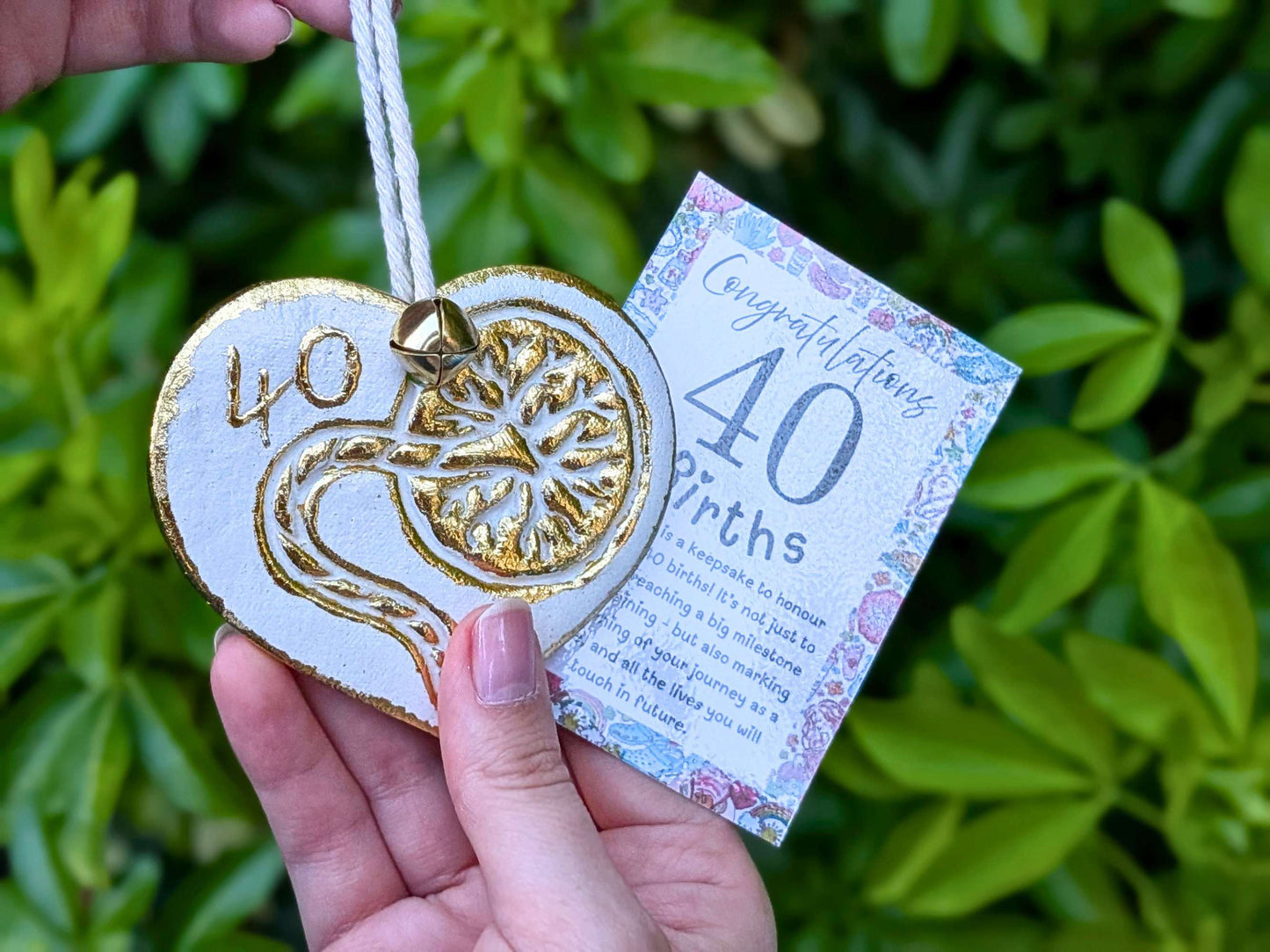 A hand holding a white heart-shaped wooden 40th birth student midwife ornament with gold foil detailing, next to a floral congratulations card, with a blurred green garden background.
