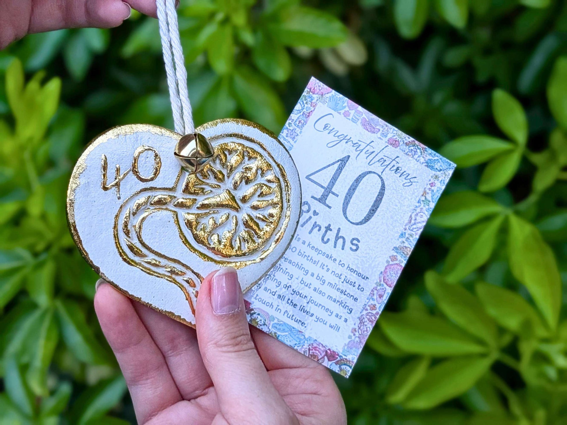 A hand holding a white heart-shaped wooden 40th birth student midwife ornament with gold foil detailing, next to a floral congratulations card, with a blurred green garden background.