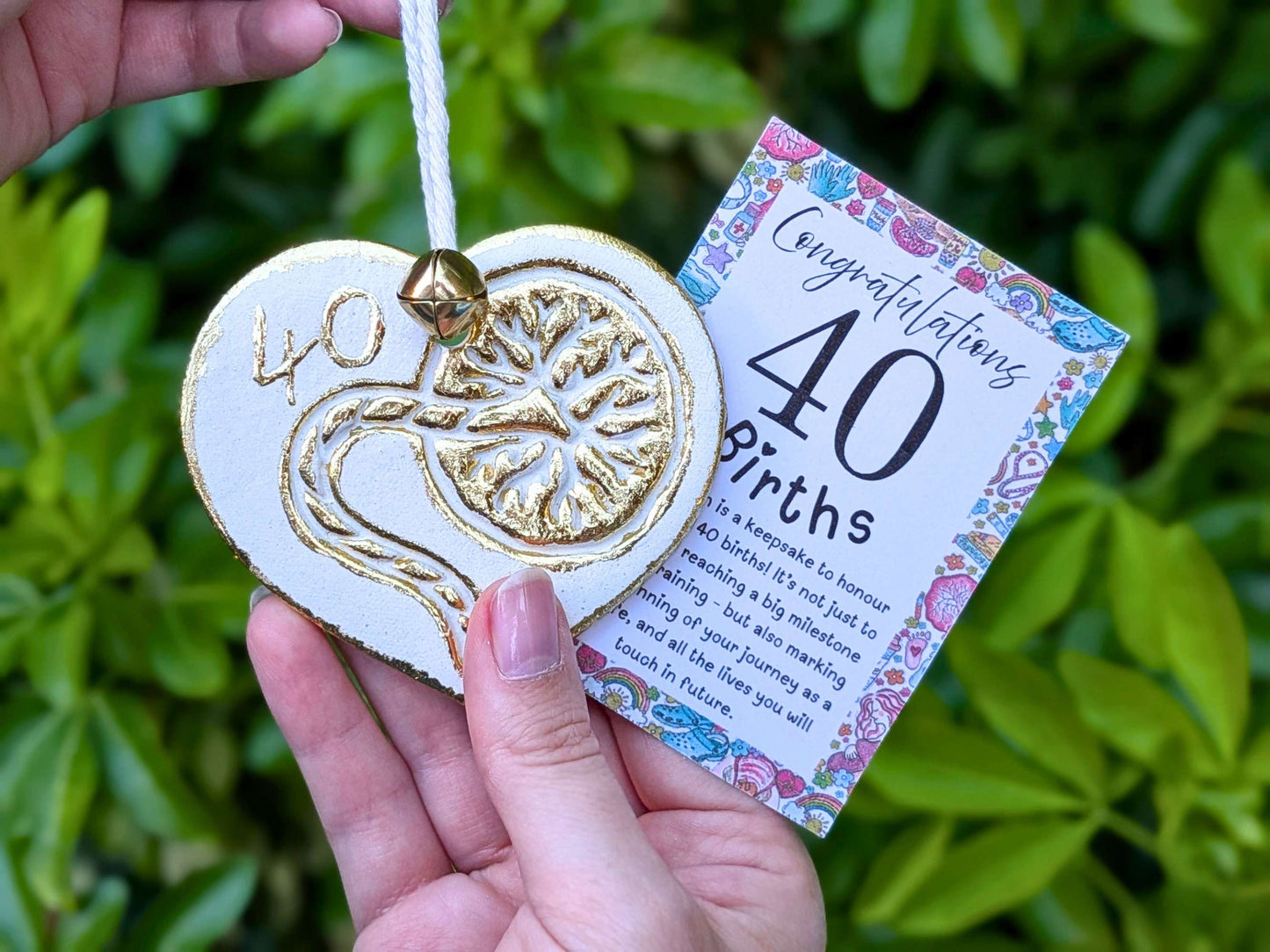 A hand holding a white heart-shaped wooden 40th birth student midwife ornament with gold foil detailing, next to a floral congratulations card, with a blurred green garden background.