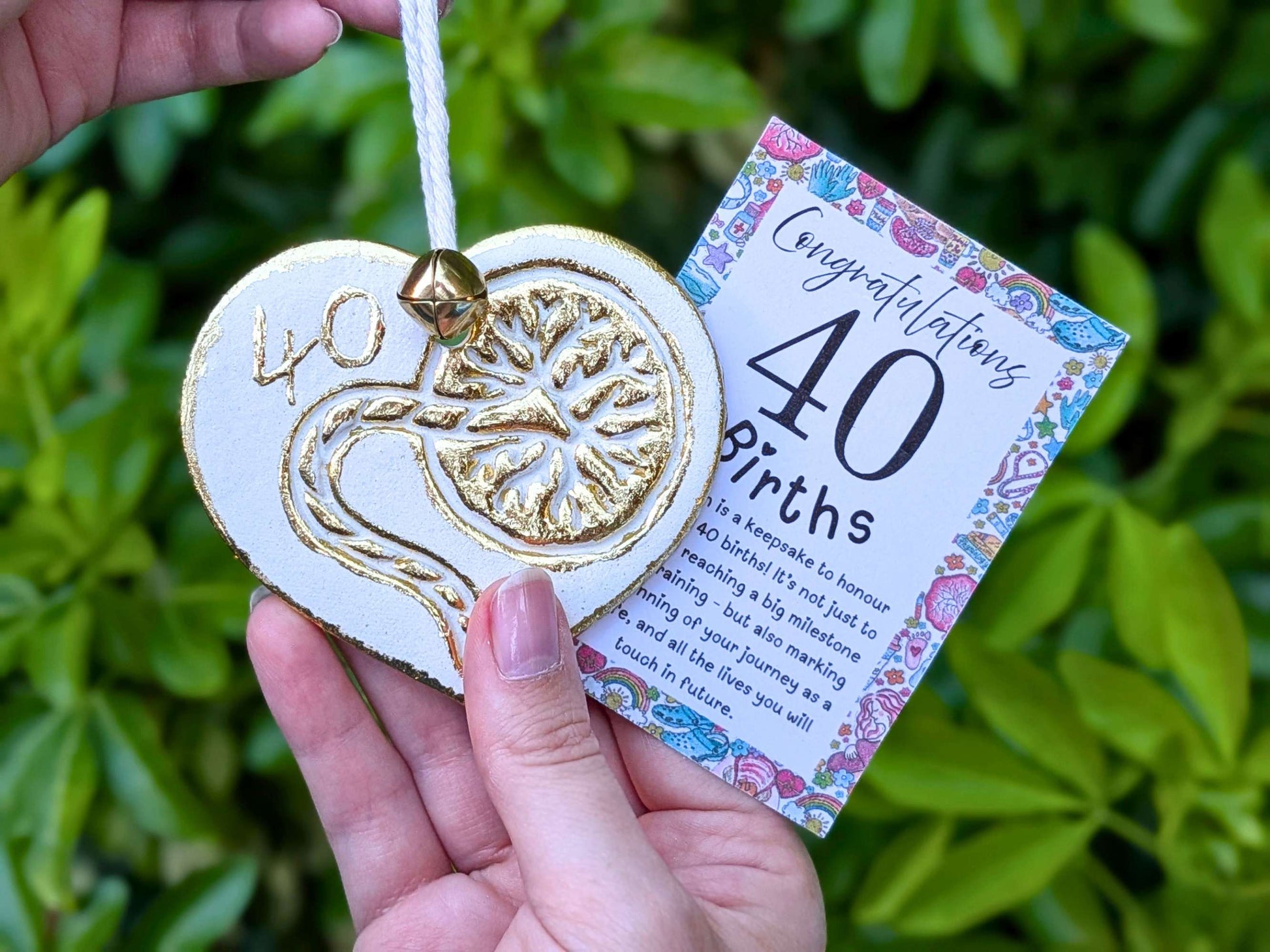 A hand holding a white heart-shaped wooden 40th birth student midwife ornament with gold foil detailing, next to a floral congratulations card, with a blurred green garden background.