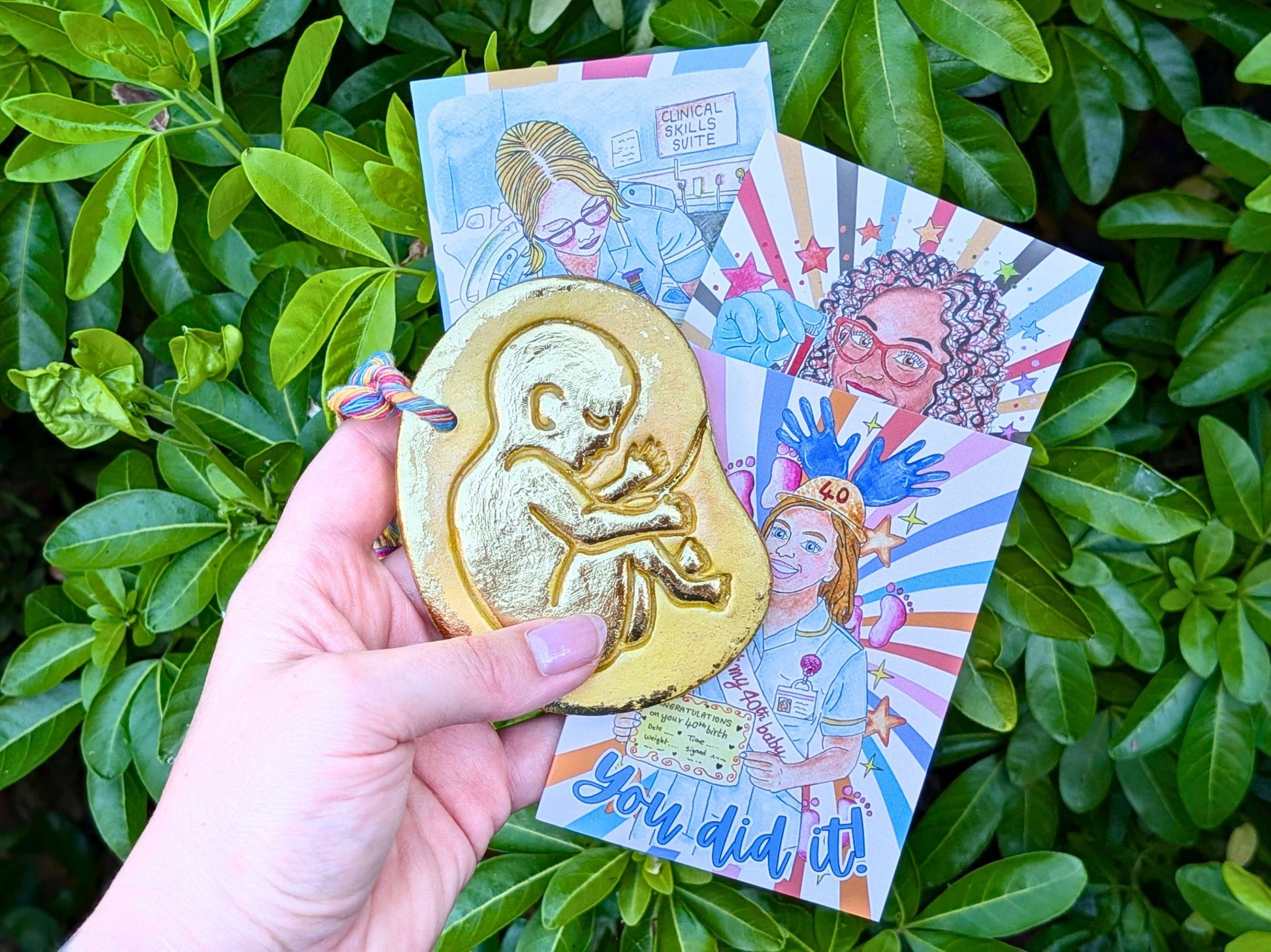 A hand holds a gold-colored baby in the womb ornament featuring an embossed fetus design in front of colorful illustrated cards, set against a background of green leaves.