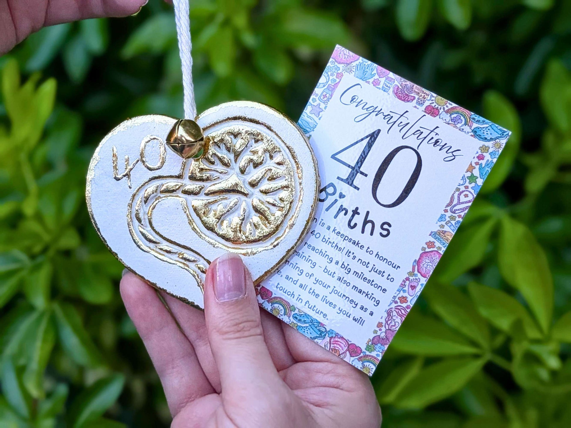 A hand holding a white heart-shaped wooden 40th birth student midwife ornament with gold foil detailing, next to a floral congratulations card, with a blurred green garden background.