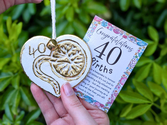 A hand holding a white heart-shaped wooden 40th birth student midwife ornament with gold foil detailing, next to a floral congratulations card, with a blurred green garden background.