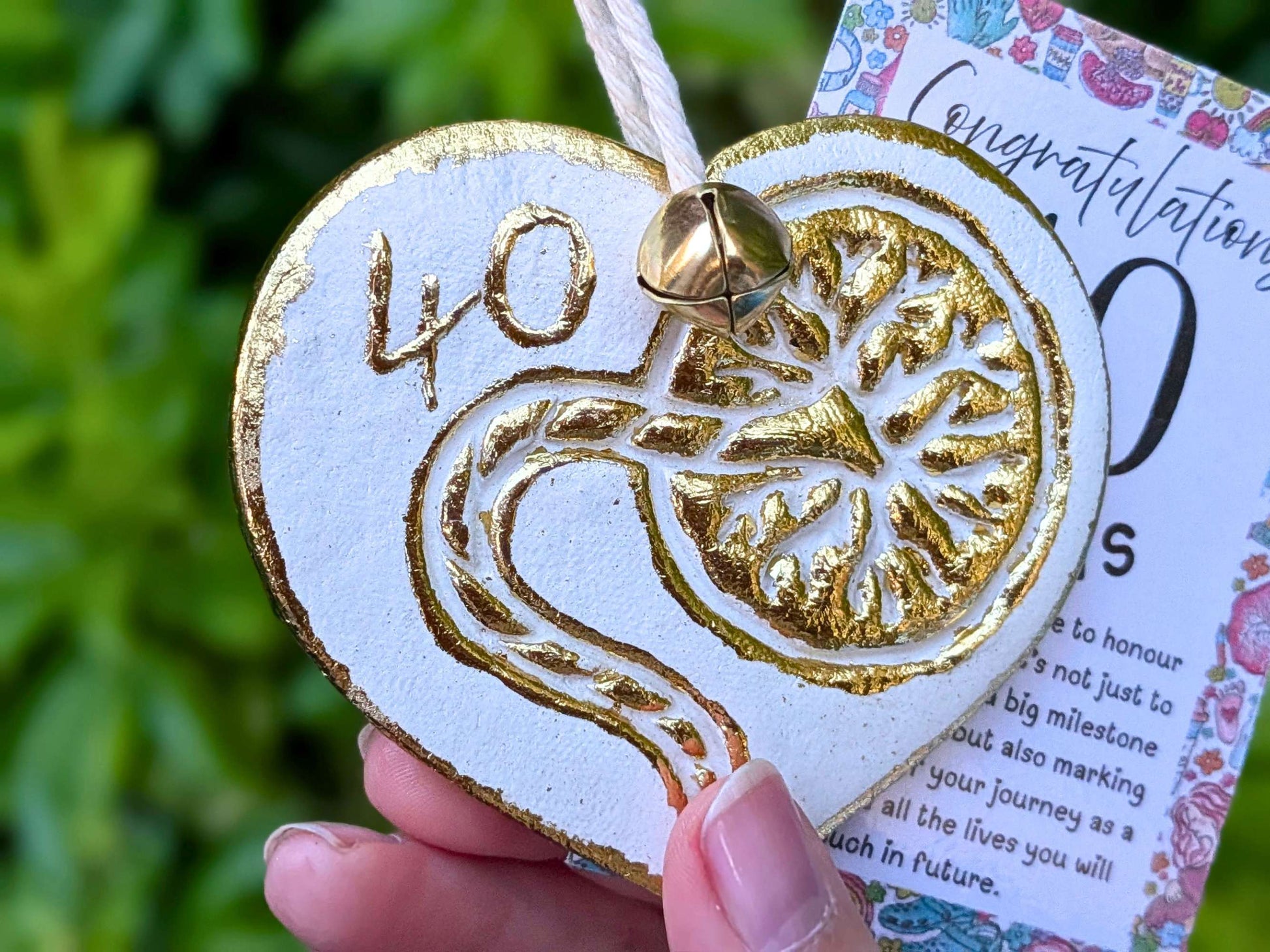 A hand holding a white heart-shaped wooden 40th birthday ornament with gold foil detailing, next to a floral congratulations card, with a blurred green garden background.