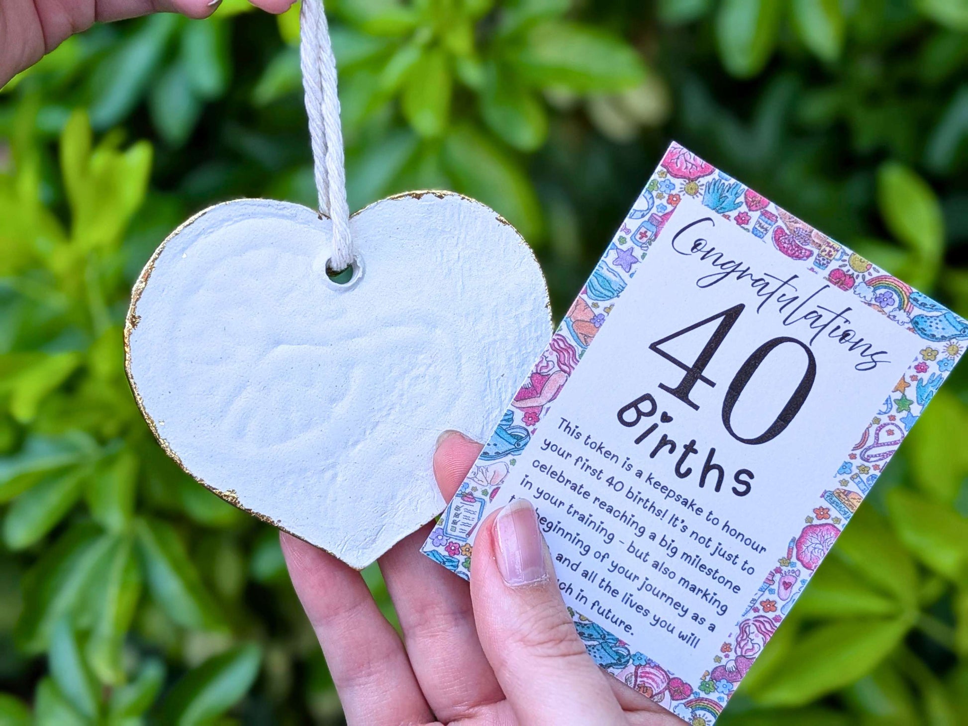 A hand holds a white heart-shaped wooden keepsake token with a hanging loop next to a colorful 'Congratulations 40th Births' card, set against a blurred green garden background.