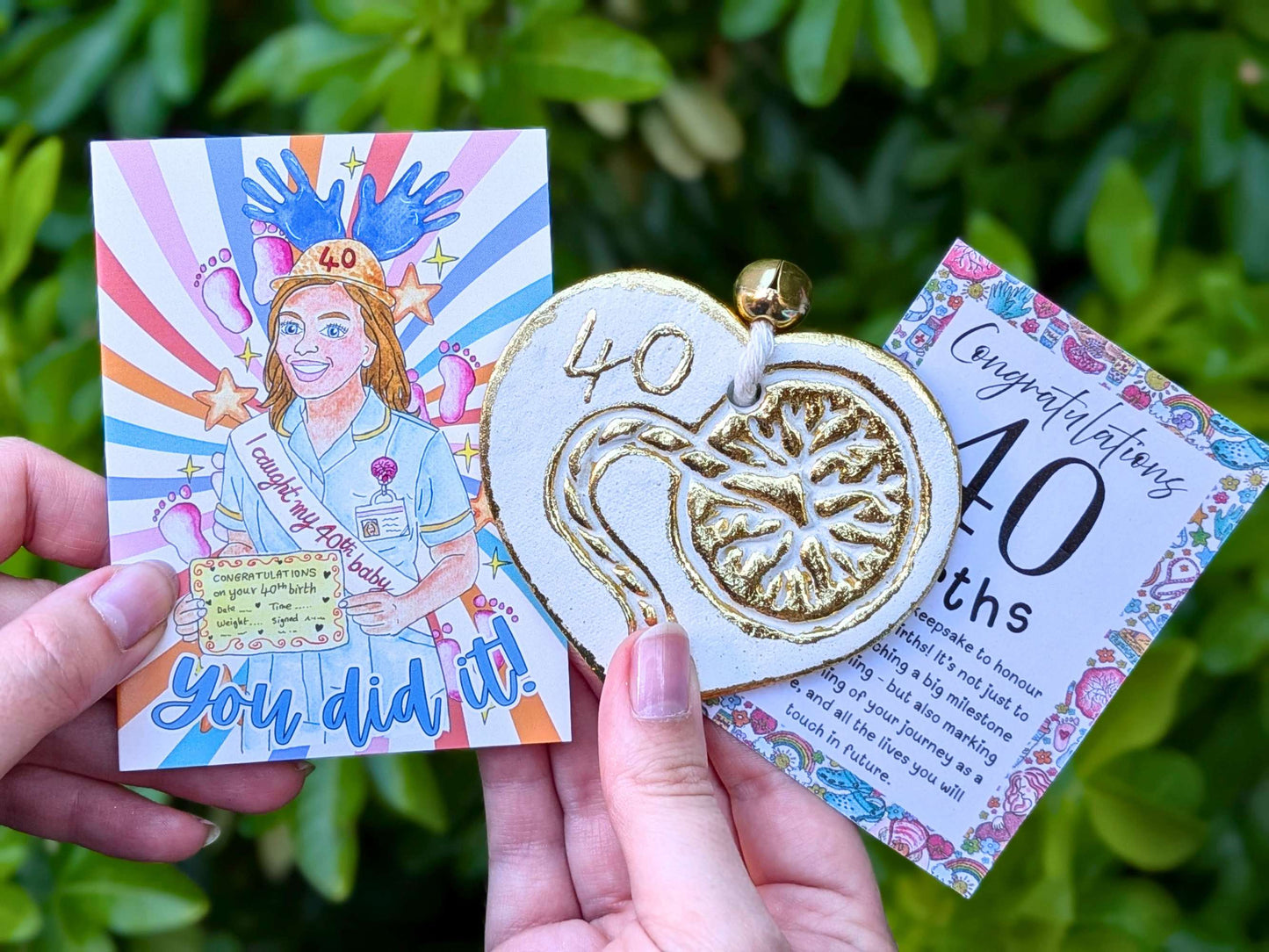 A person holds a 40th birthday gift set outdoors against a green leaf background. The set includes a square illustrated card with a colorful midwife illustration and the text 'You did it!', a gold and white heart-shaped pendant with '40' embossed, and a floral 'Congratulations 40th births' message card.