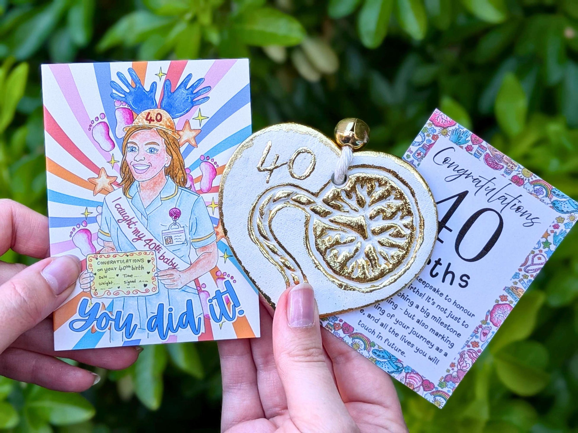 A person holds a 40th birthday gift set outdoors against a green leaf background. The set includes a square illustrated card with a colorful midwife illustration and the text 'You did it!', a gold and white heart-shaped pendant with '40' embossed, and a floral 'Congratulations 40th births' message card.