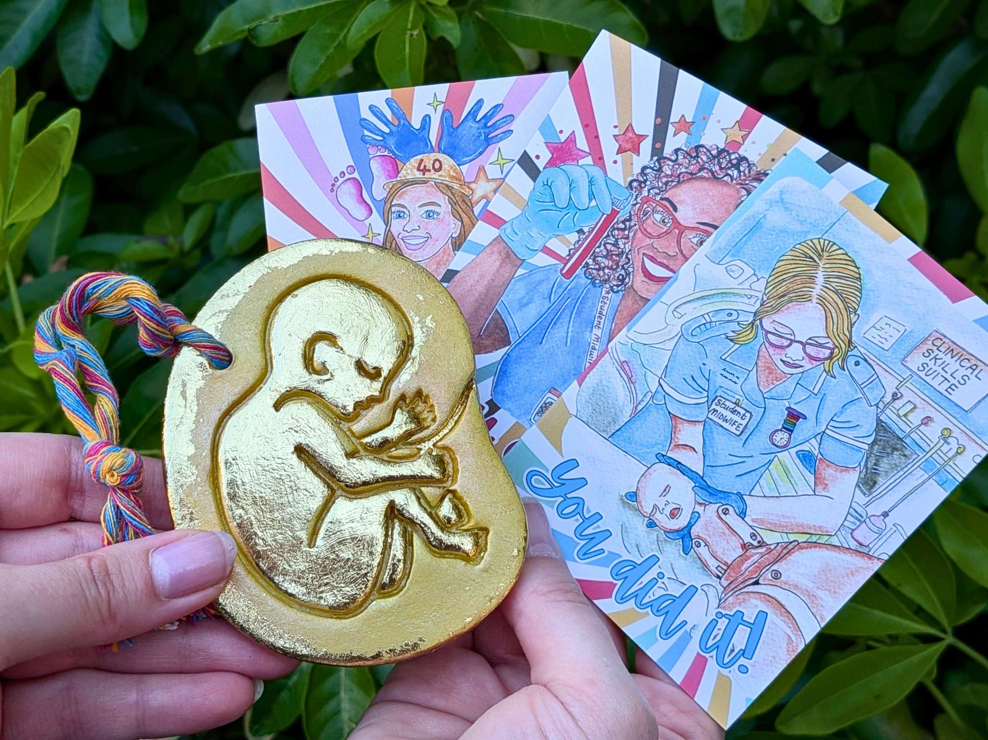 A hand holds a gold-colored baby in the womb ornament featuring an embossed fetus design in front of colorful illustrated cards, set against a background of green leaves.