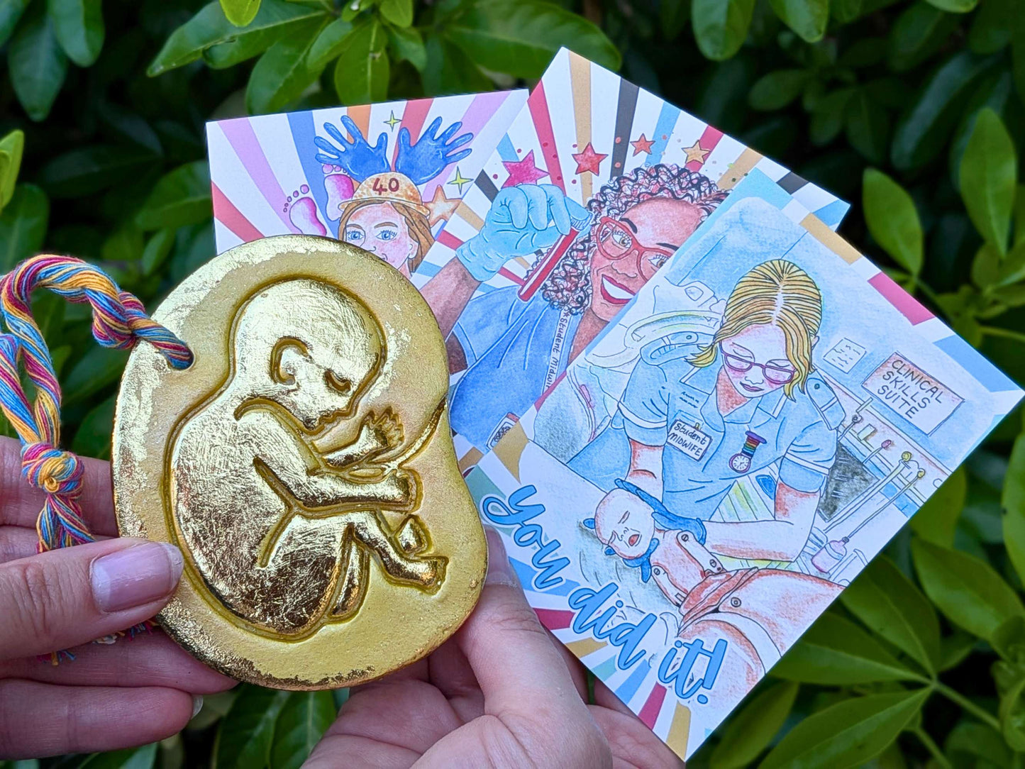 A hand holds a gold-colored baby in the womb ornament featuring an embossed fetus design in front of colorful illustrated cards, set against a background of green leaves.