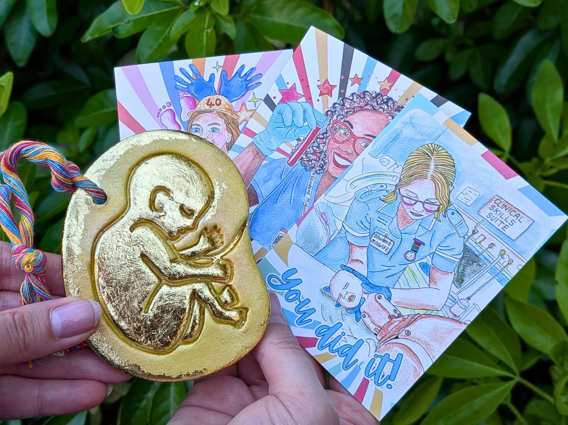 A hand holds a gold-colored baby in the womb ornament featuring an embossed fetus design in front of colorful illustrated cards, set against a background of green leaves.
