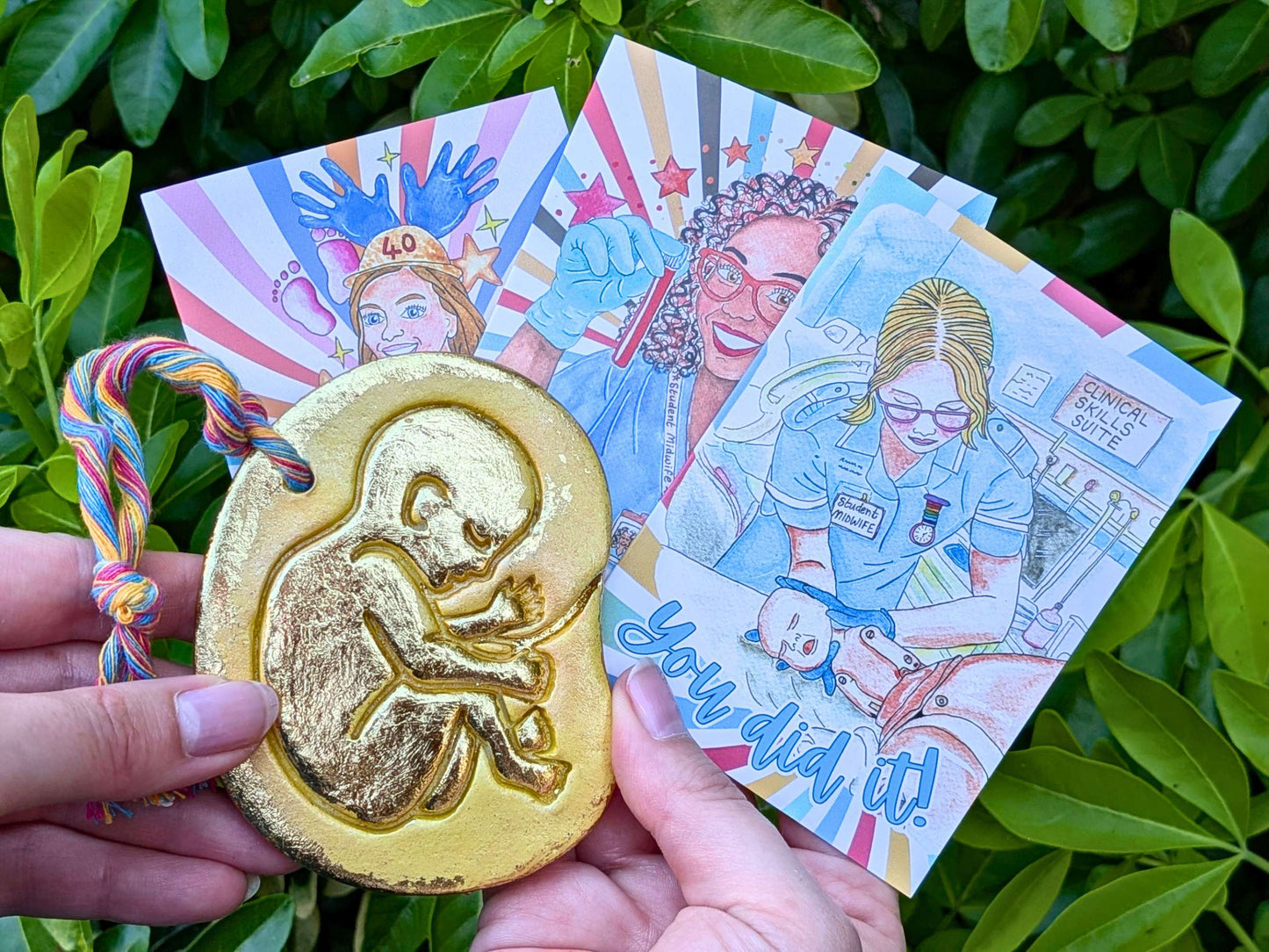 A hand holds a gold-colored baby in the womb ornament featuring an embossed fetus design in front of colorful illustrated cards, set against a background of green leaves.