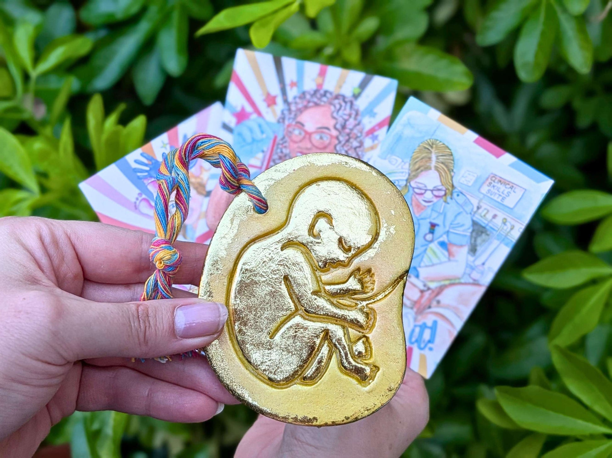 A hand holds a gold-colored baby in the womb ornament featuring an embossed fetus design in front of colorful illustrated cards, set against a background of green leaves.