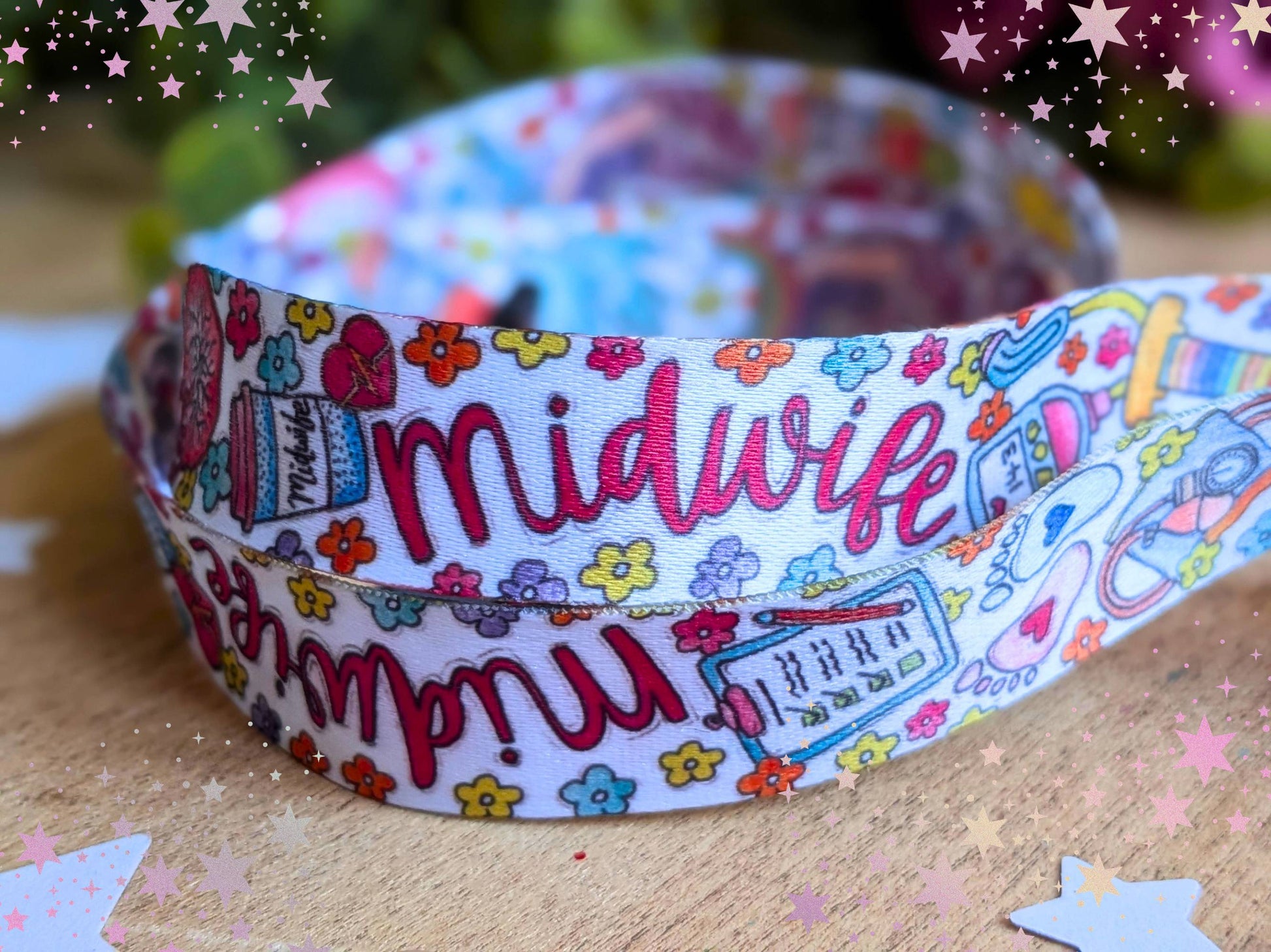 A coiled midwife lanyard featuring colorful hand-drawn illustrations and the word 'midwife' in pink script, displayed on a wooden surface with decorative star overlays.