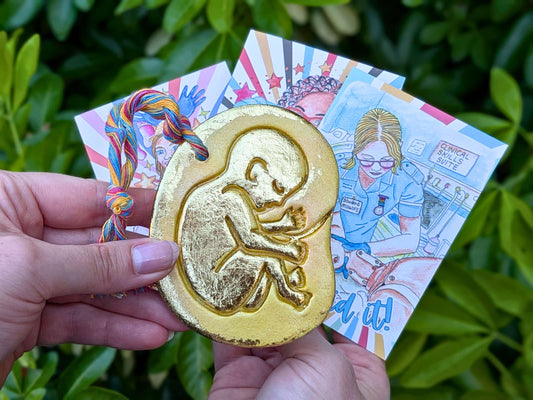 A hand holds a gold-colored baby in the womb ornament featuring an embossed fetus design in front of colorful illustrated cards, set against a background of green leaves.