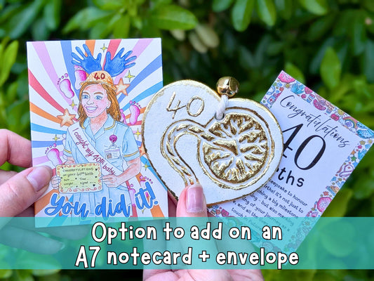A person's hands hold a 40th birth card set against a background of green foliage. The set includes a colorful illustrated card, a gold and white enamel heart-shaped pendant with '40' and a snake motif, and a floral 'Congratulations on your 40th birth' card.