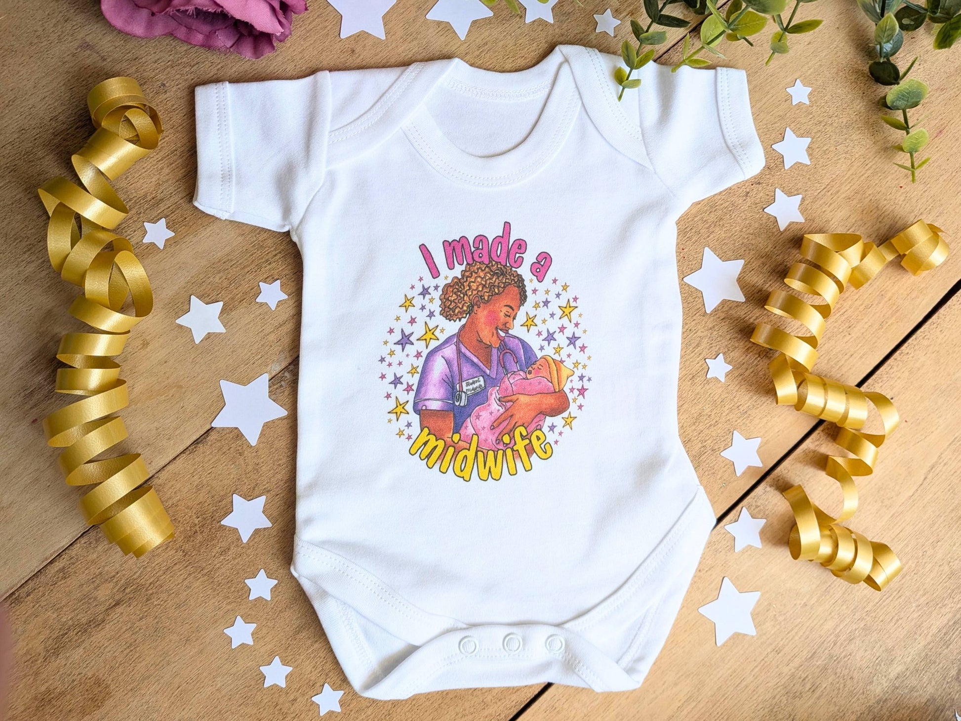 A white short-sleeve baby bodysuit laid flat on a wooden surface, decorated with gold ribbon curls, white star cutouts, and greenery. The bodysuit features a colorful graphic of a midwife holding a baby with the text 'I made a midwife'.