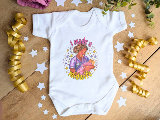 A white short-sleeve baby bodysuit laid flat on a wooden surface, decorated with gold ribbon curls, white star cutouts, and greenery. The bodysuit features a colorful graphic of a midwife holding a baby with the text 'I made a midwife'.